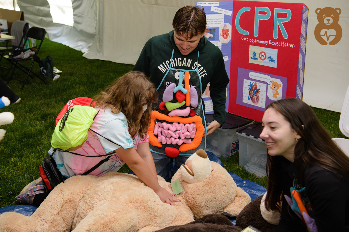 A child learning about CPR from a medical student on a large stuffed bear.